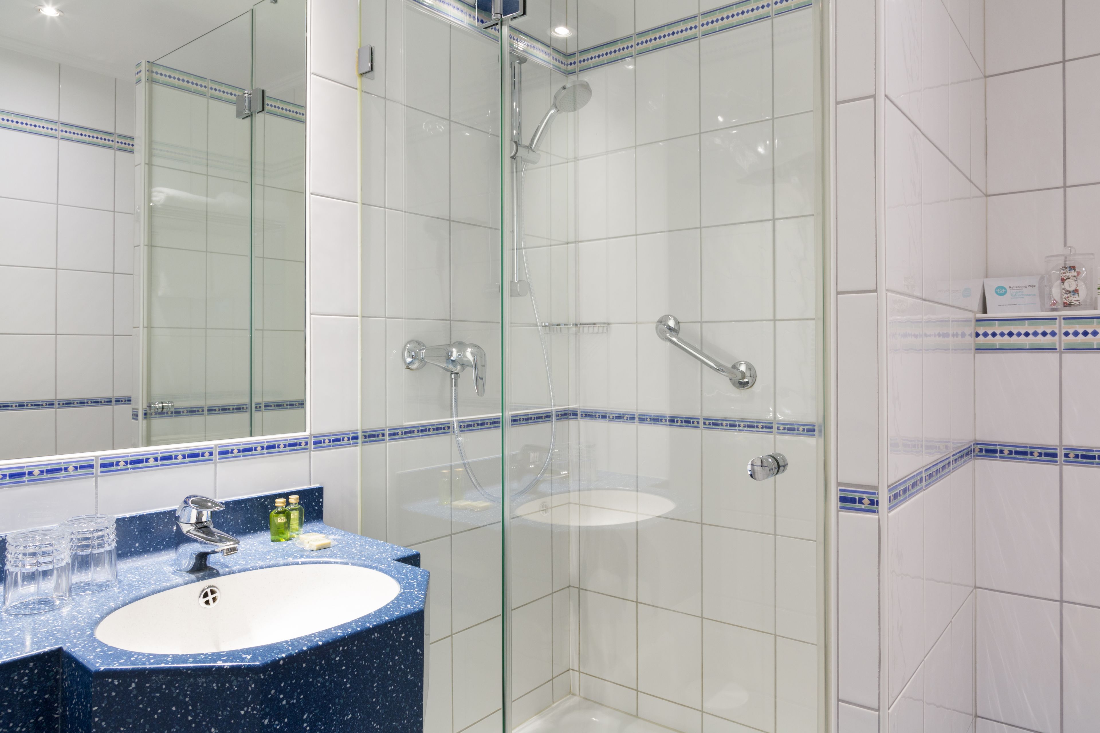 Un baño con ducha envuelta en vidrio, un lavabo con encimera azul y paredes de azulejos blancos con detalles en azul.