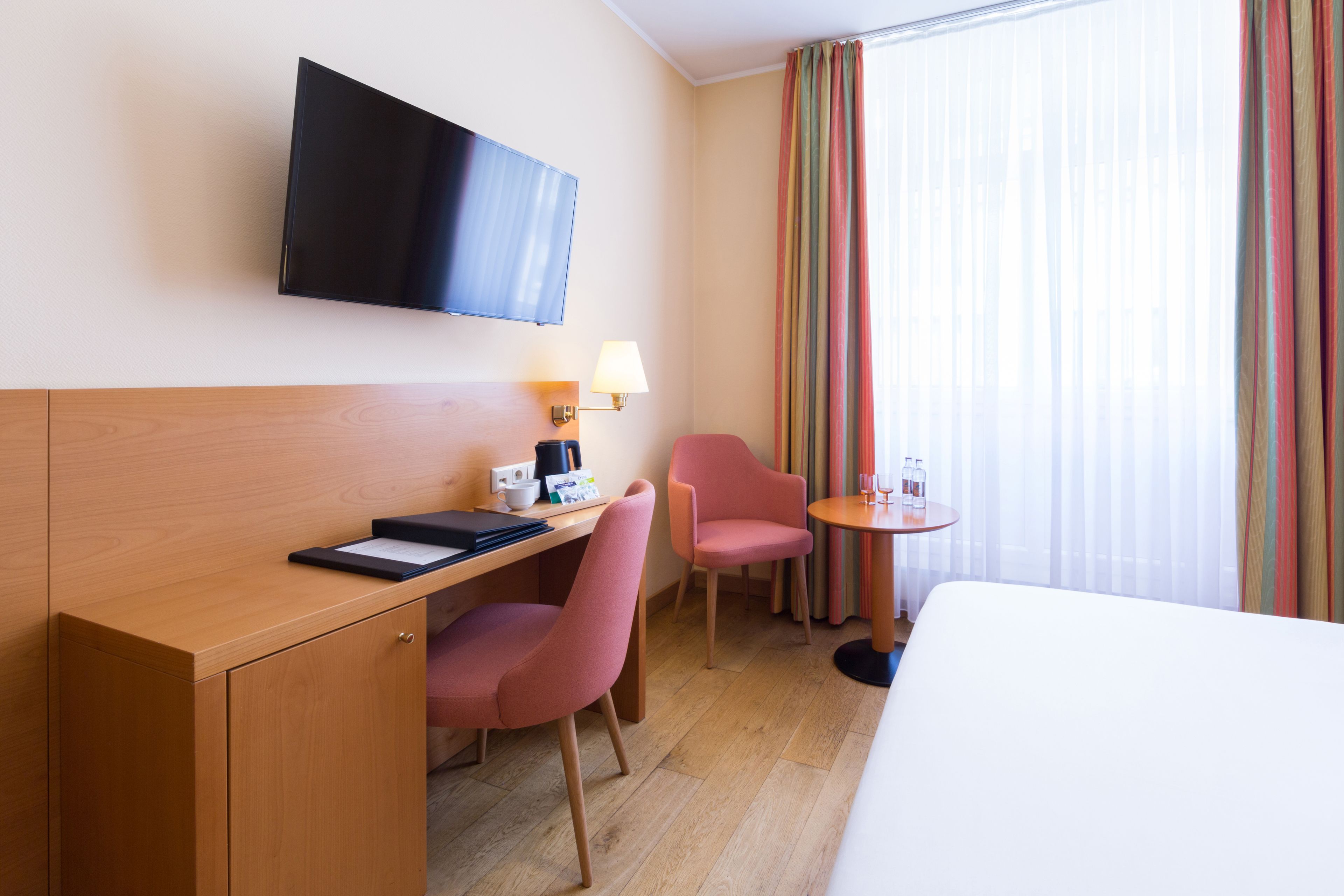 A hotel room with a wooden desk, a pink chair, a small round table with a lamp, a television mounted on the wall, and a bed.