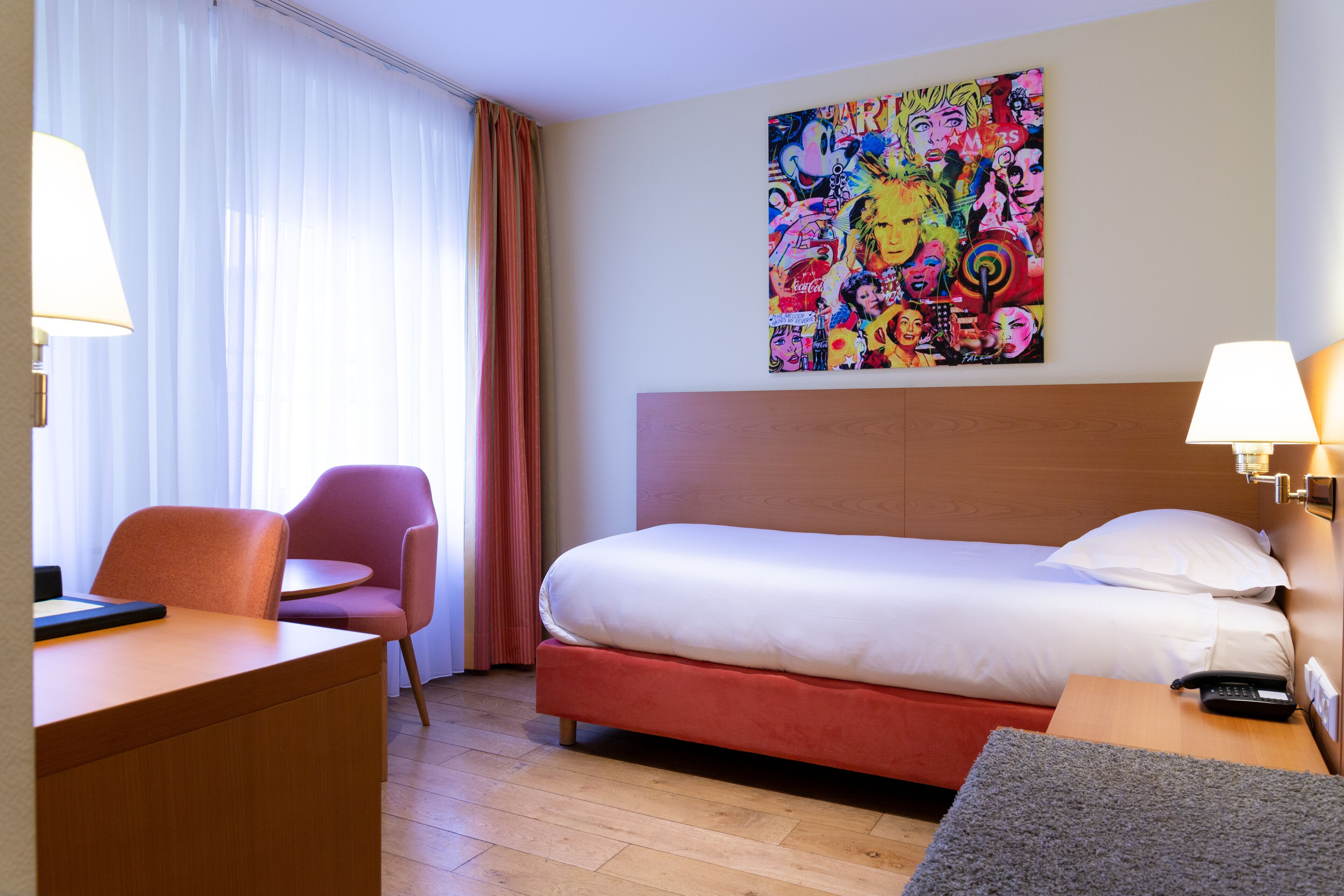 A hotel room with a bed, a desk, a chair, a colorful artwork on the wall, and a lamp on the bedside table.
