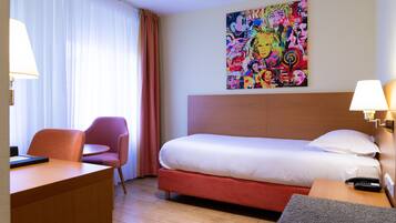 A hotel room with a bed, a desk, a chair, a colorful artwork on the wall, and a lamp on the bedside table.