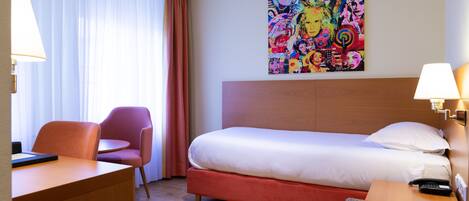 A hotel room with a bed, a desk, a chair, a colorful artwork on the wall, and a lamp on the bedside table.