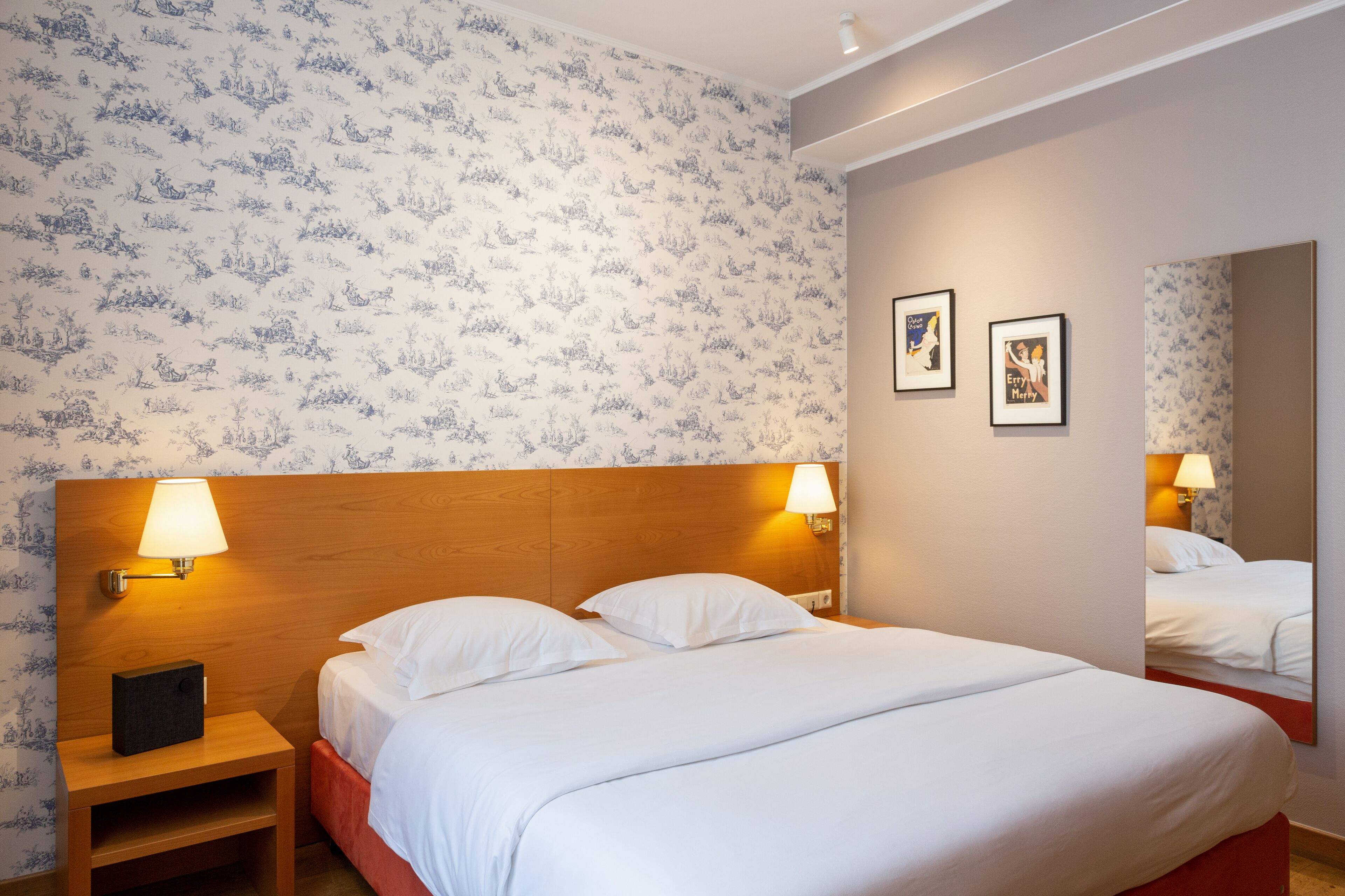 A hotel room with a wooden headboard, two bedside lamps, a bed with white linens, a nightstand with a lamp, and a mirror on the wall.