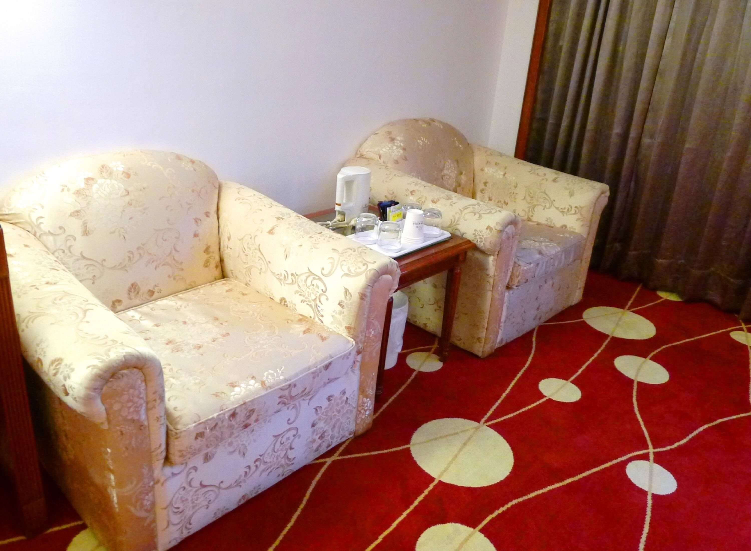 Habitación de hotel con dos sillones, una mesita con bandeja y una alfombra roja con un diseño circular.