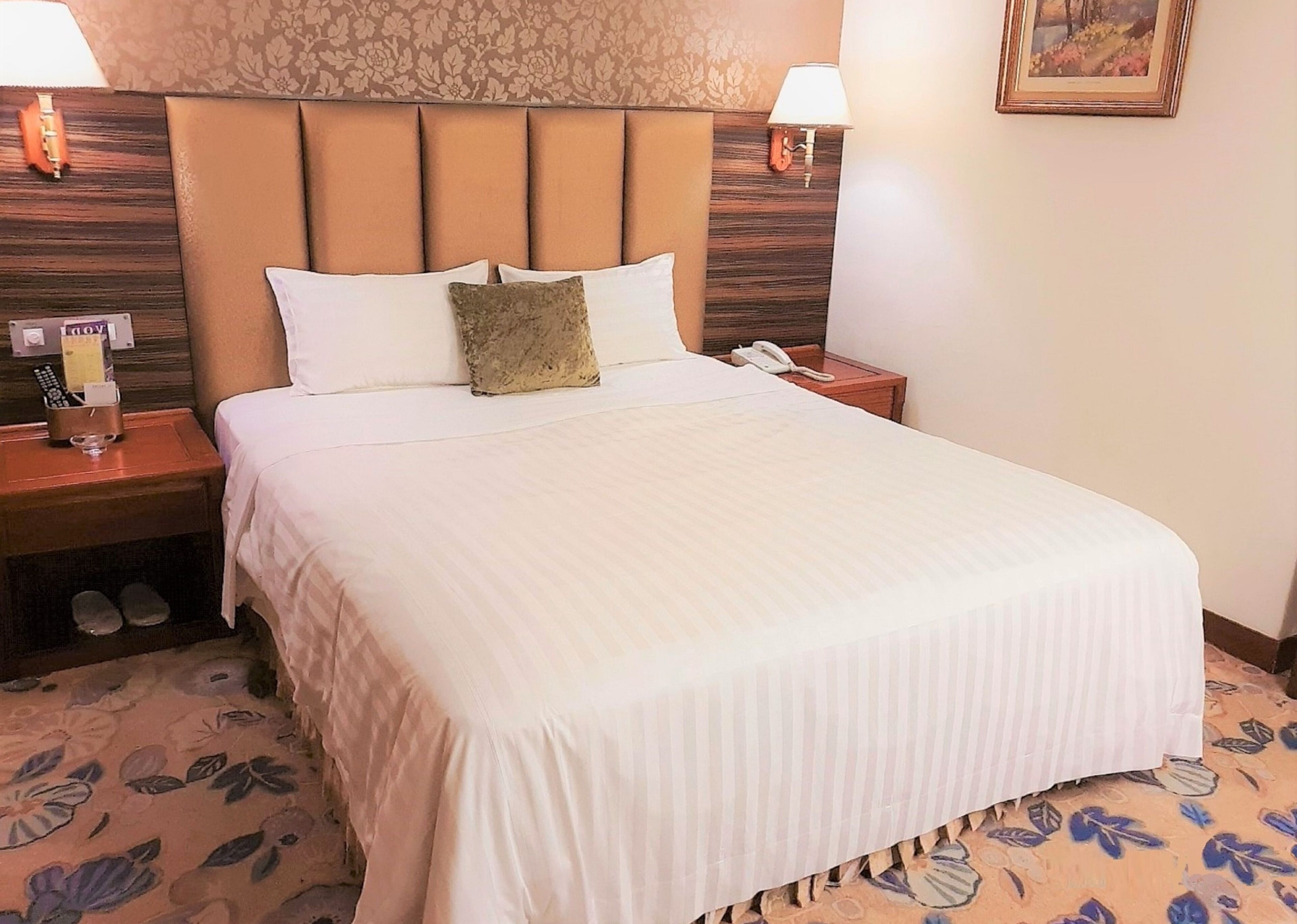 A hotel room with a large bed, two bedside tables, a nightstand, a lamp, a phone, and a floral-patterned carpet.