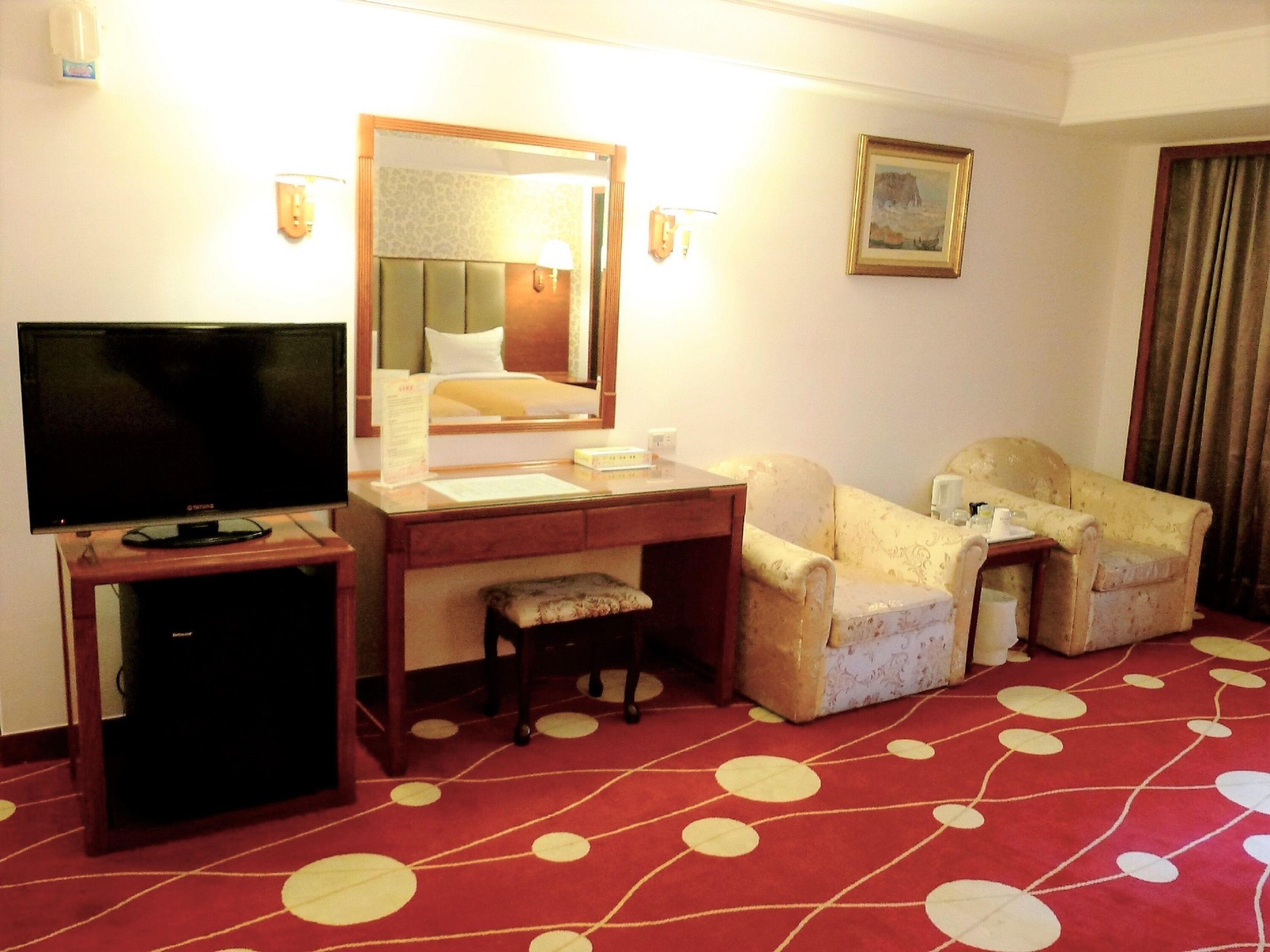 A hotel room with a television, a desk, a chair, a small stool, a mirror, a framed picture on the wall, and a red carpet with white polka dots.