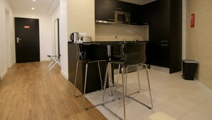 A modern kitchen with a microwave, toaster, and sink. There are two bar stools and a small table. The floor is wooden, and the walls are tiled.