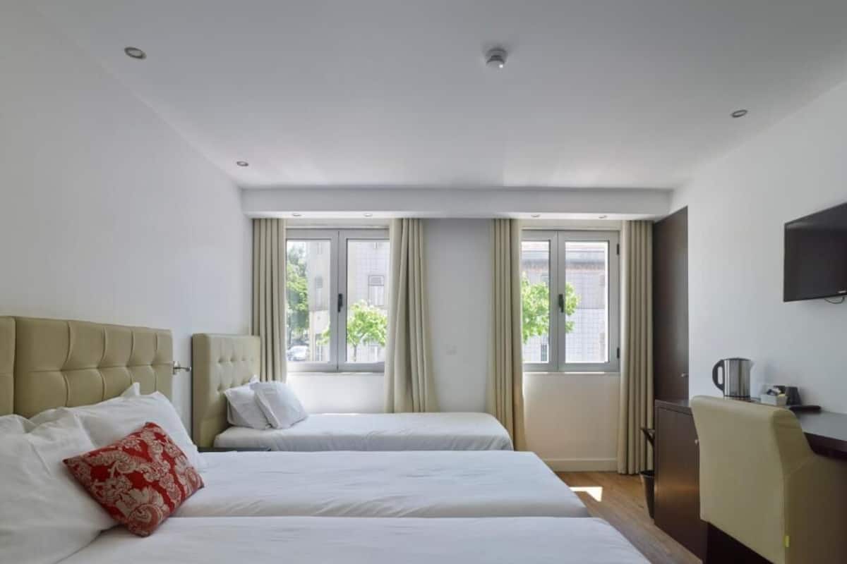 A hotel room with two beds, a desk, a television, and large windows with curtains.