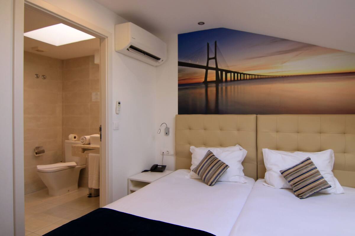 A hotel room with a large bed, a shower, a toilet, and a painting of a bridge on the wall.