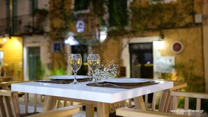 Une table dressée pour deux personnes, avec des verres à vin et une petite composition florale décorative, le tout mis en valeur par l’arrière-plan d’un bâtiment recouvert de lierre et orné d’une horloge.