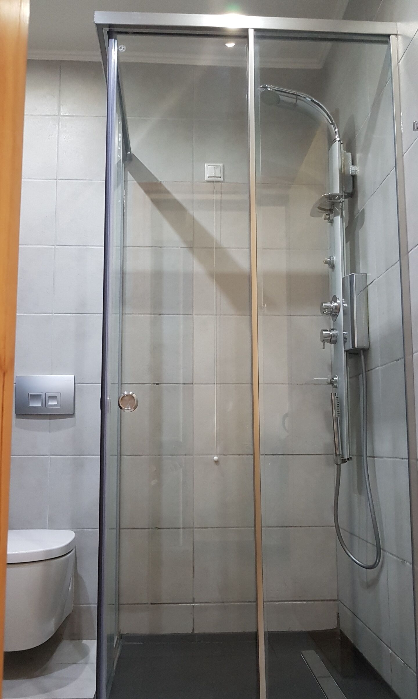 A glass-enclosed shower stall with a handheld showerhead, a wall-mounted faucet, and a control panel with two buttons.