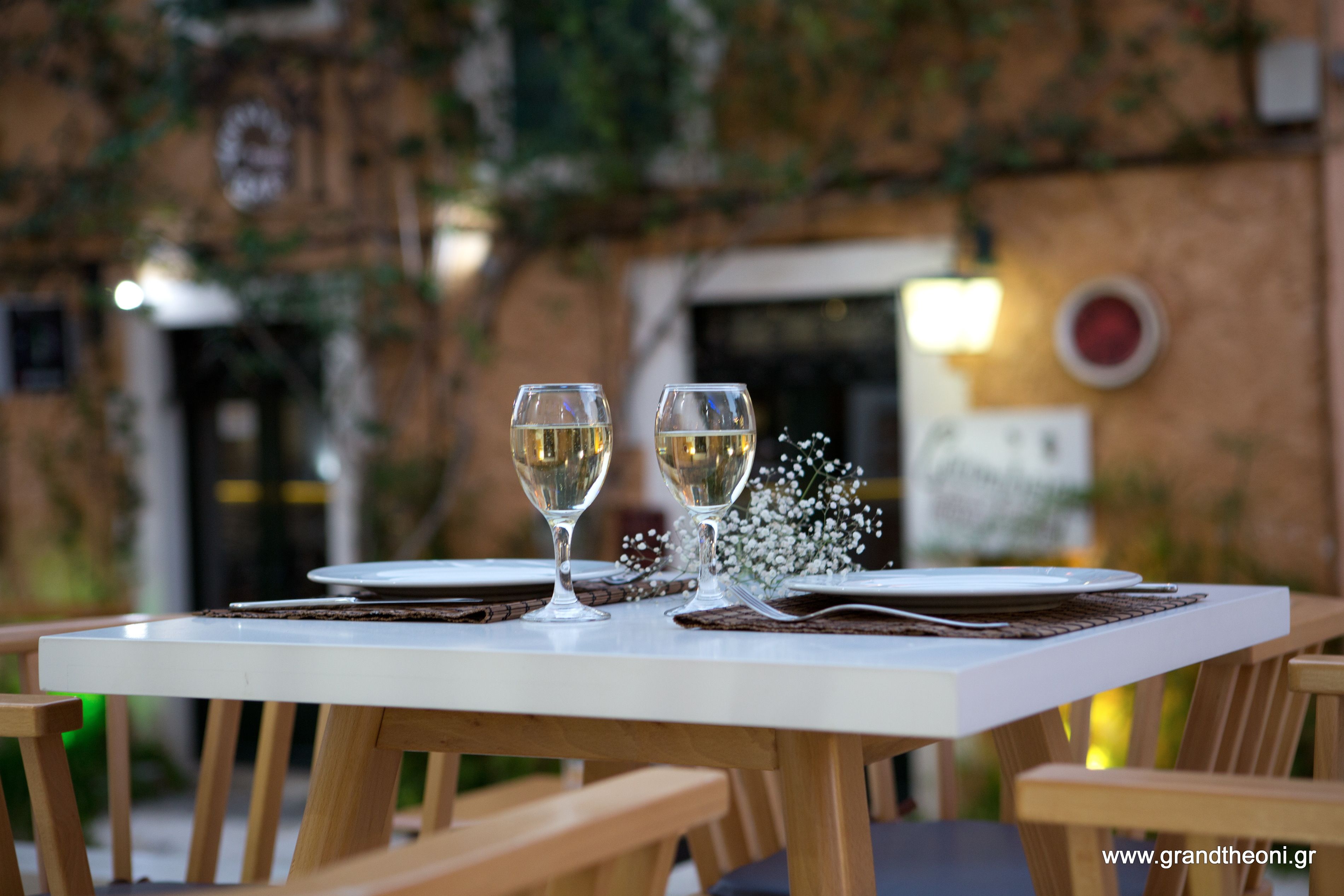 A table set for two with wine glasses and plates, surrounded by wooden chairs.