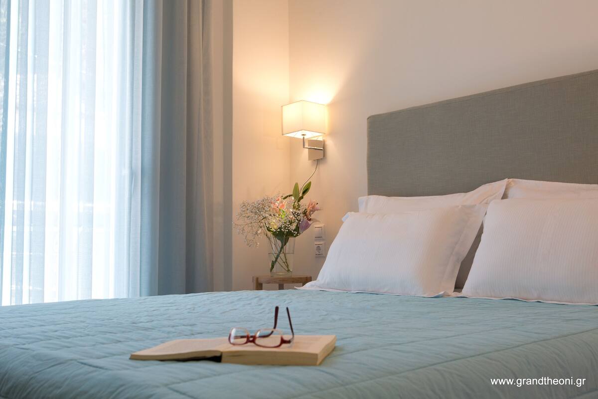 Un lit bien fait, avec des oreillers blancs et une couverture bleue, une table de chevet ornée d’un vase de fleurs et une lampe fixée au mur.