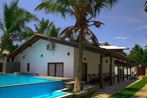 A poolside area with a swimming pool, a covered patio with chairs, and a palm tree.