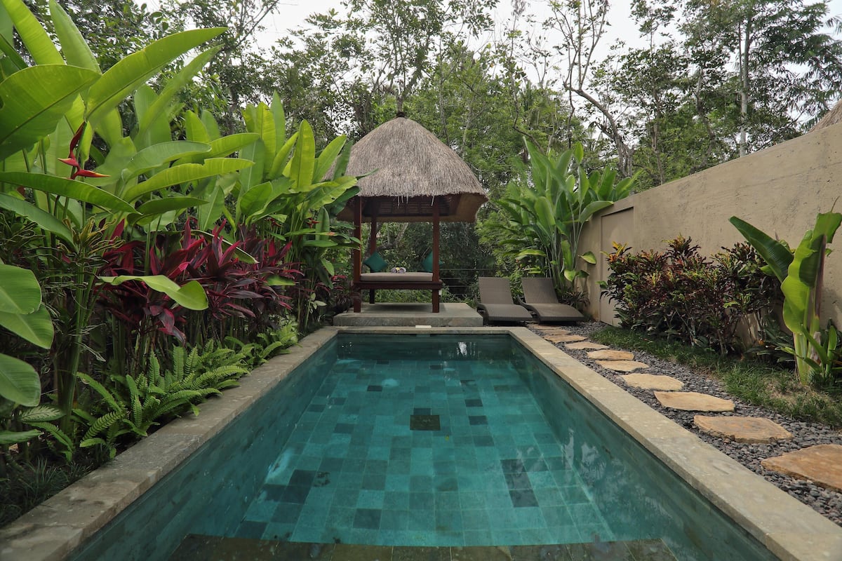 Una piscina rectangular con un pabellón de techo de paja en el centro, rodeada de exuberante follaje y un borde de piedra.