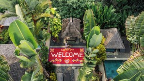 Ein tropisches Resort mit einem strohgedeckten Gebäude, einer Fußmatte mit floralen Mustern und einem Pool, der von üppiger Vegetation umgeben ist.