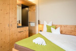 Habitación de hotel con armario de madera, cama con ropa de cama verde, almohadas blancas y una toalla doblada con cuidado.