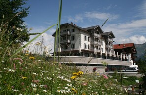 Un edificio de varios pisos con balcones, un área de restaurante con mesas al aire libre, y montañas al fondo.