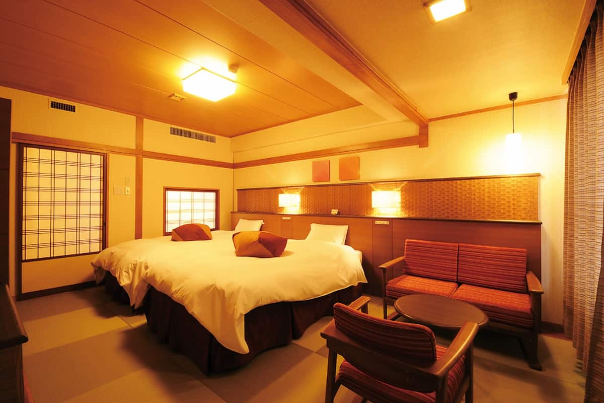 A traditional Japanese-style hotel room with tatami flooring, a large bed with pillows, a low table, and a red cushioned bench.