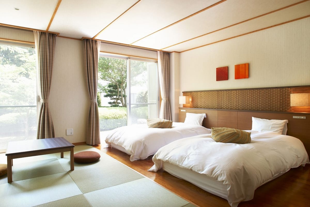 A hotel room with two beds, a small table, and a view of greenery through the window.
