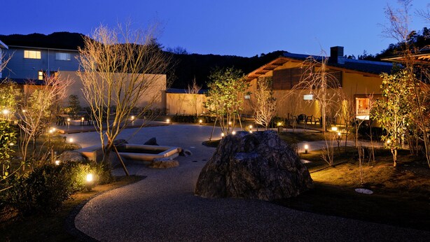 A well-lit garden area with pathways, a large rock, and a building with a tiled roof. - Satoyama no Kyujitsu Kyoto Keburikawa (Kameoka)