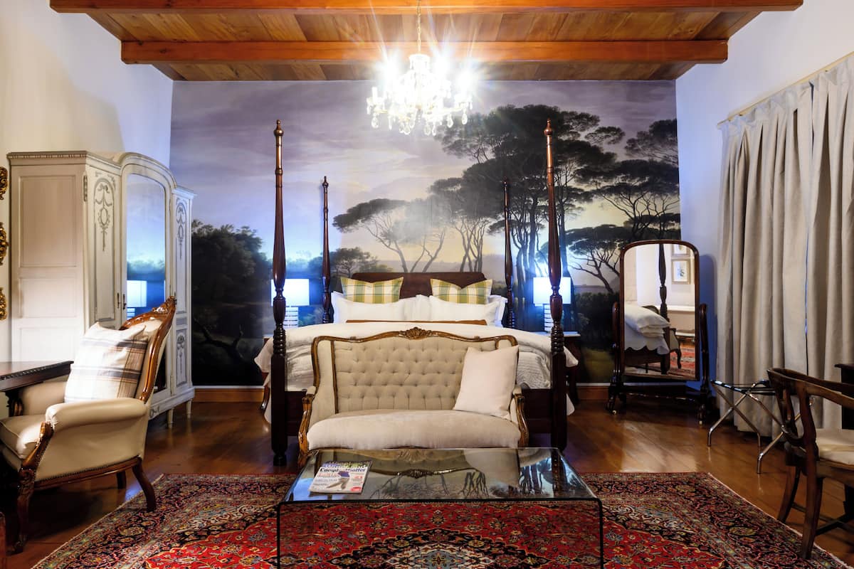 Ein Schlafzimmer mit einem großen Bett, einer Kristallleuchter, einem Spiegel und Blick auf Bäume durch ein Fenster.