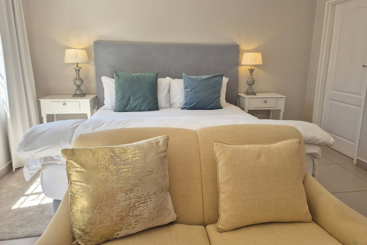 Ein Hotelzimmer mit einem großen Bett, zwei Nachttischen mit Lampen, einem beige gefärbten Sofa mit Kissen und einer schlichten Wand.
