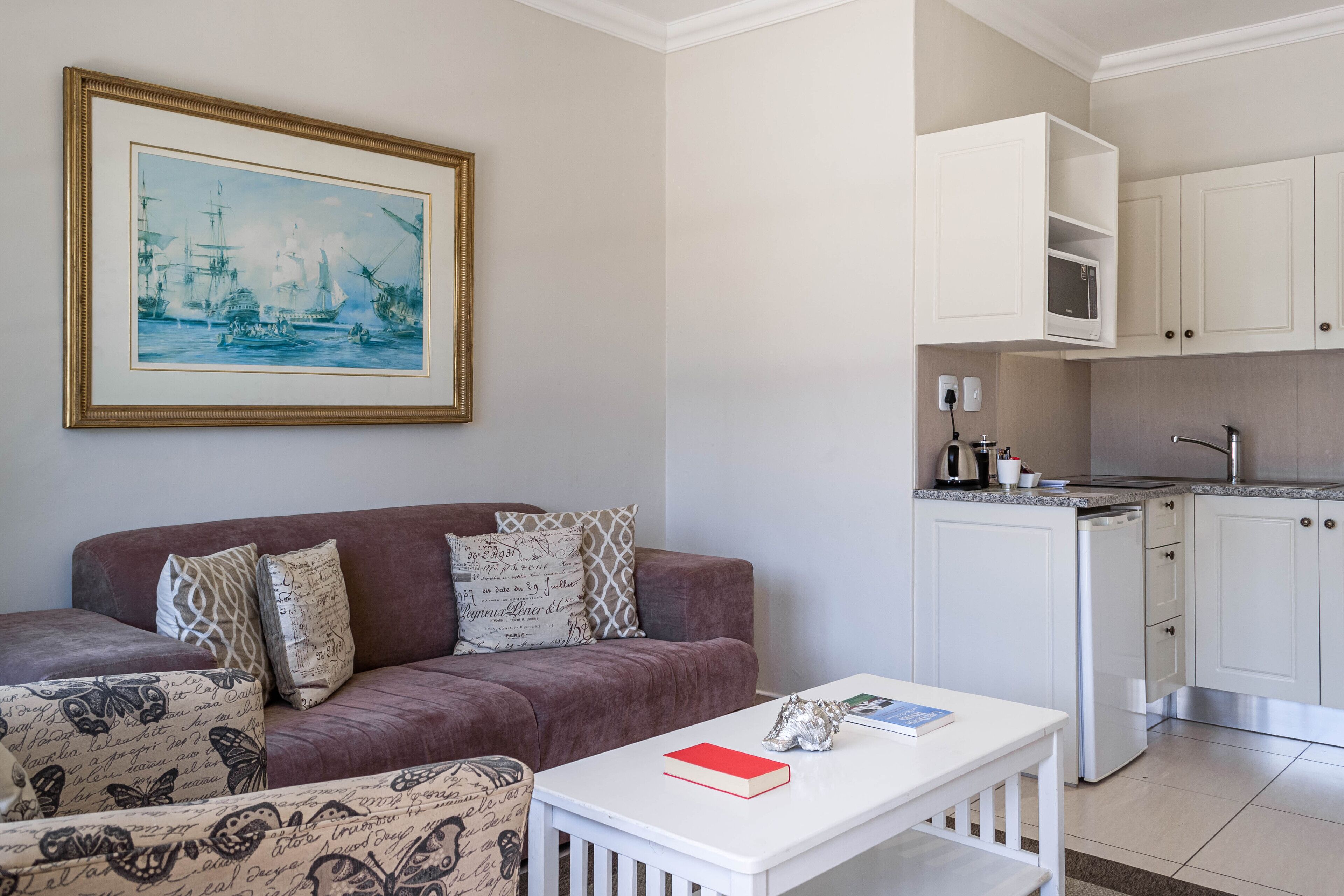 Sala de estar com sofá, mesa de centro e um quadro emoldurado. Há uma área de cozinha com armários brancos e micro-ondas.