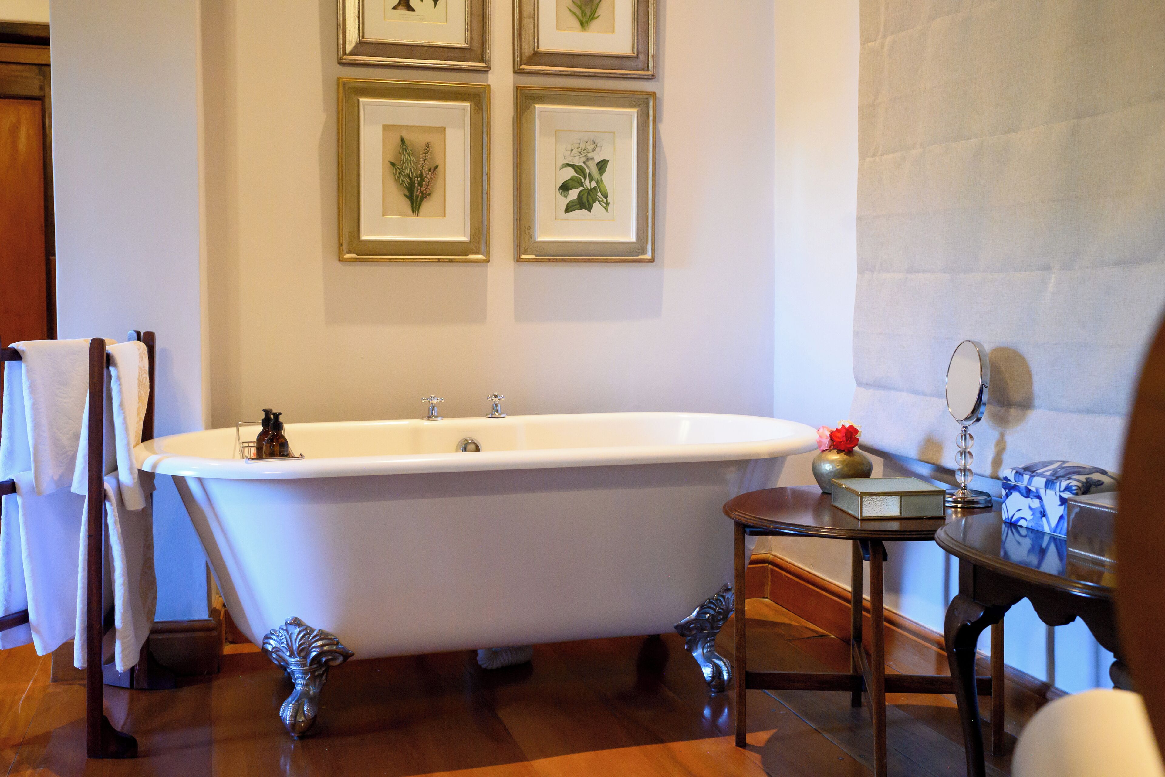 A bathroom with a freestanding bathtub, a wooden chair, a small table with a vase, and framed botanical prints on the wall.