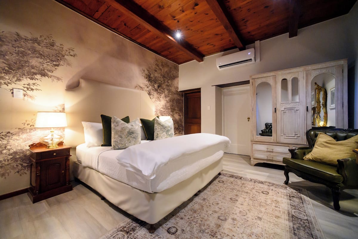 Ein Schlafzimmer mit einem großen Bett, einem Nachttisch, einer Lampe, einem grünen Sessel und einem Kleiderschrank mit Spiegel.