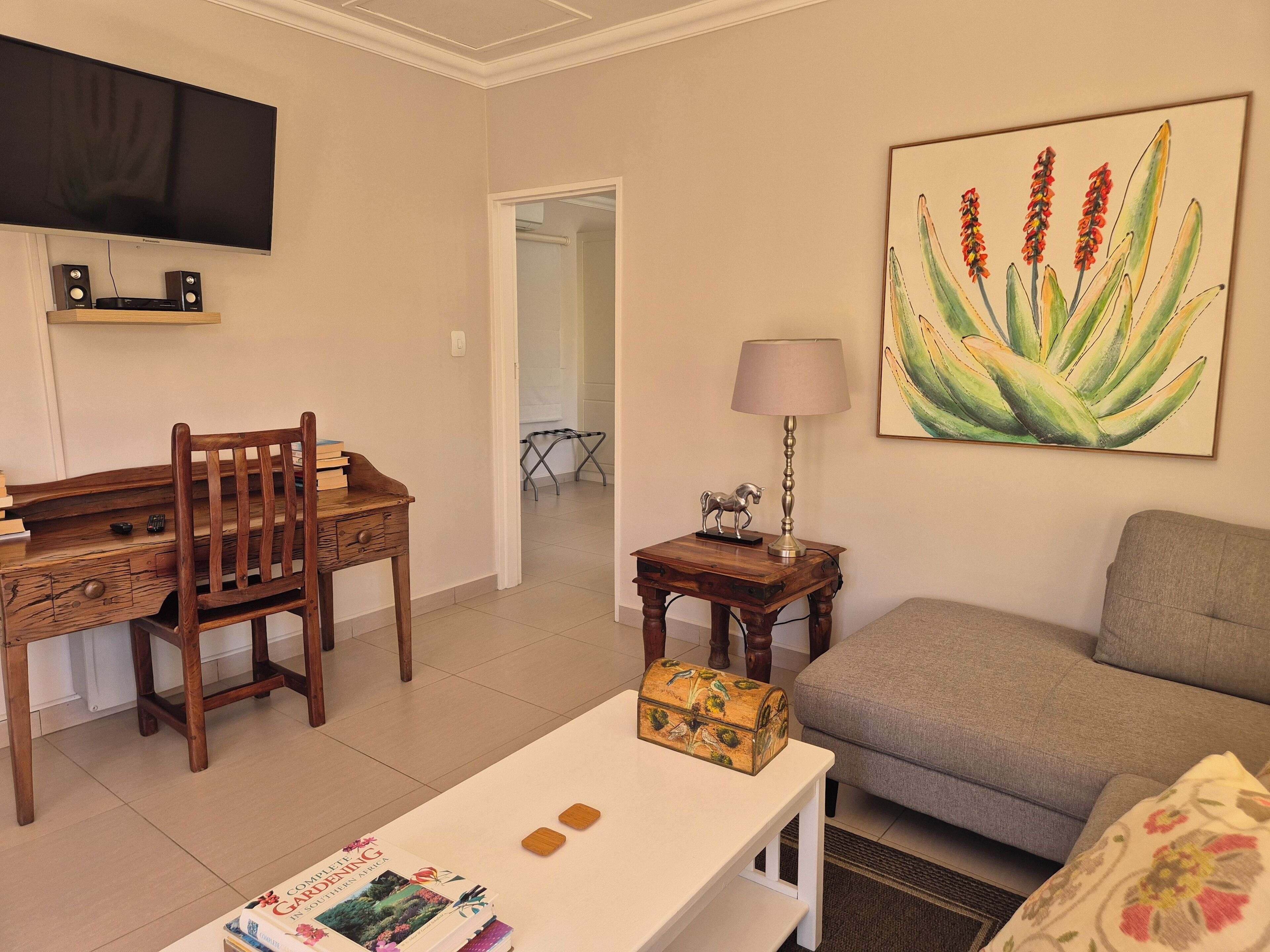 A living room with a wooden desk, a television, a sofa, and a painting of plants.
