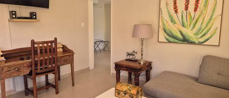 A living room with a wooden desk, a television, a sofa, and a painting of plants.