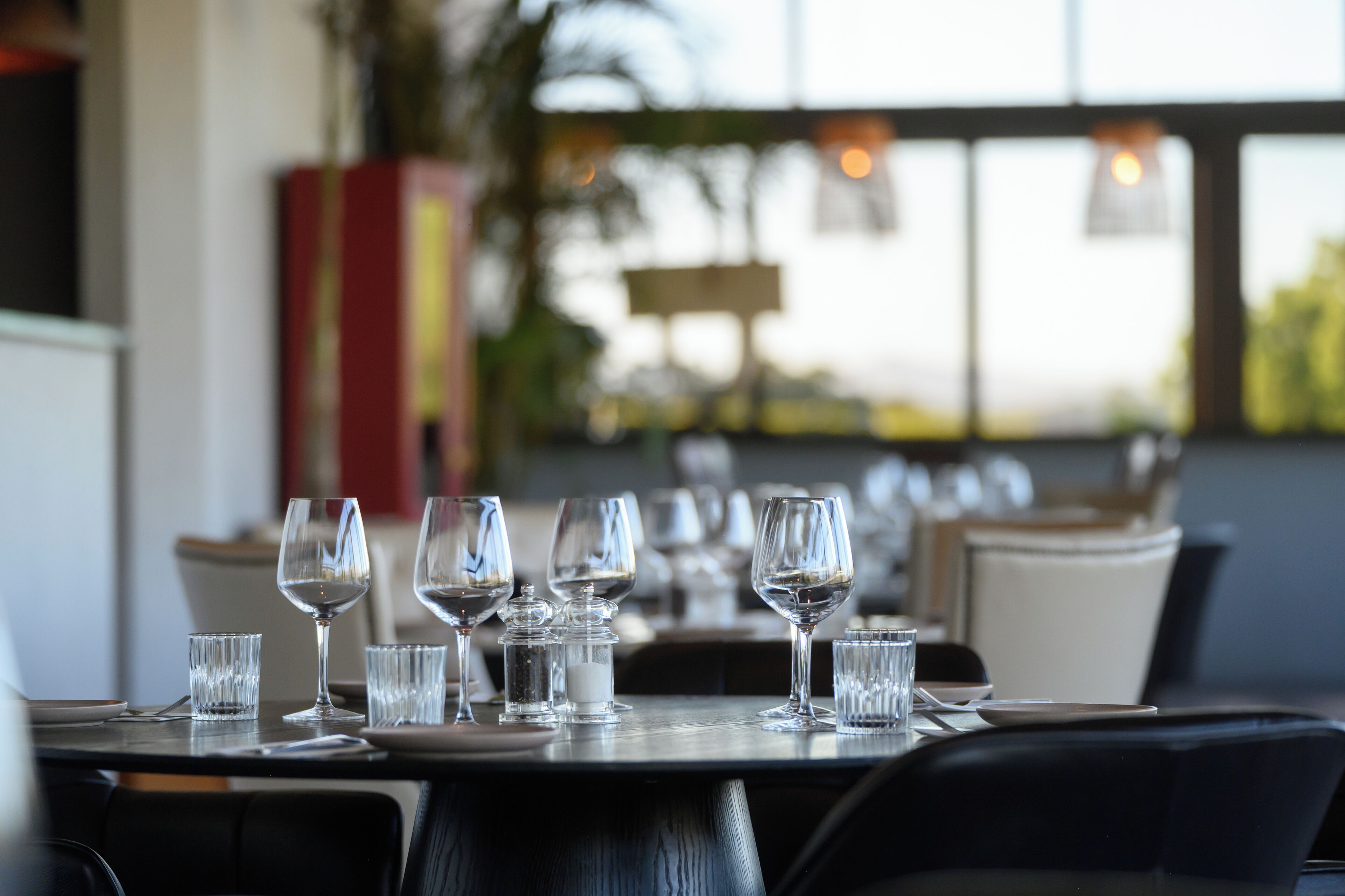 A dining table set with multiple wine glasses and water glasses, with a lamp and a plant in the background.