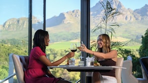 Twee personen zitten aan een tafel met uitzicht op bergen en groen.