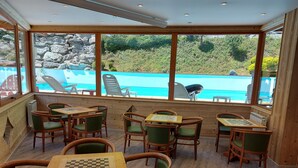 A cozy indoor seating area with wooden tables and chairs, overlooking a swimming pool and lush greenery.