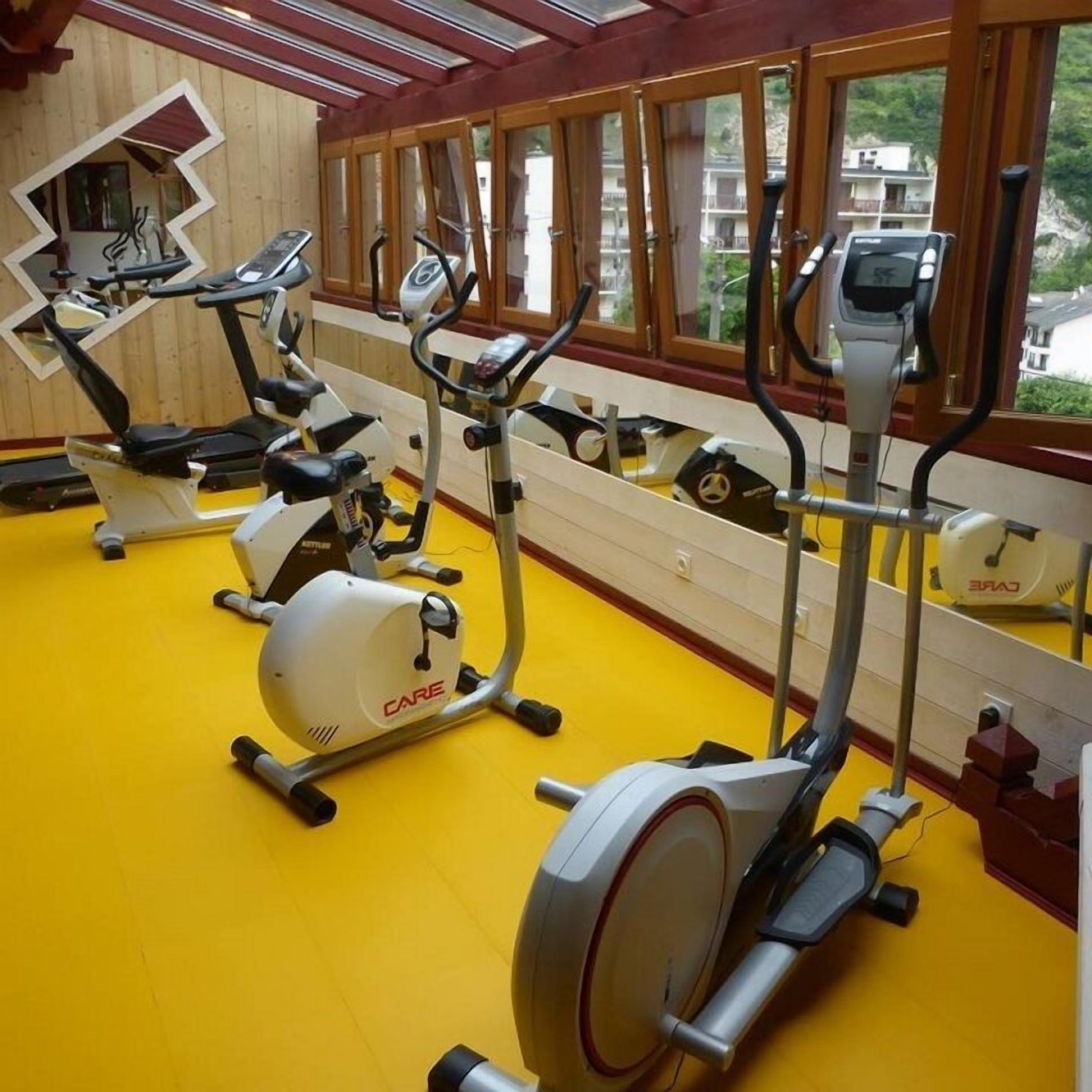 Un gimnasio con máquinas de cardio, suelo amarillo y grandes ventanales con vistas a edificios.