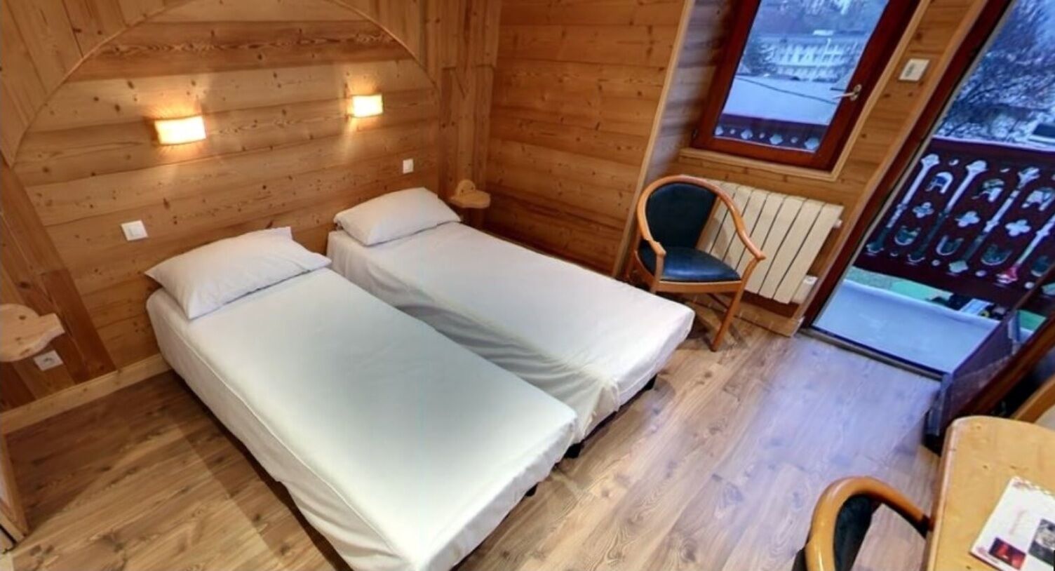 Une cabane avec deux lits simples, un plancher en bois, une chaise et une fenêtre donnant sur un paysage enneigé.