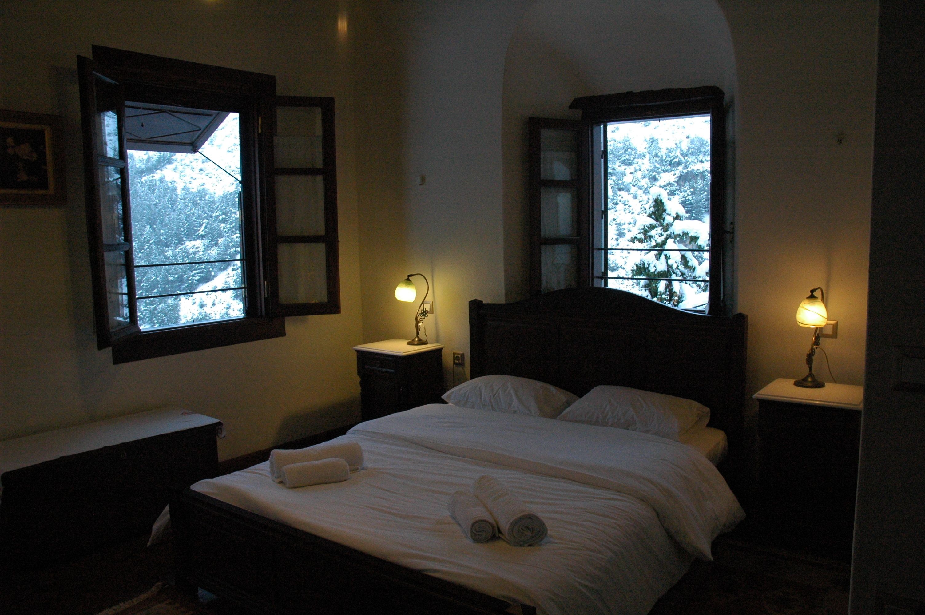 Un dormitorio con cama, dos mesitas de noche con lámparas y dos ventanas con vistas a árboles nevados.
