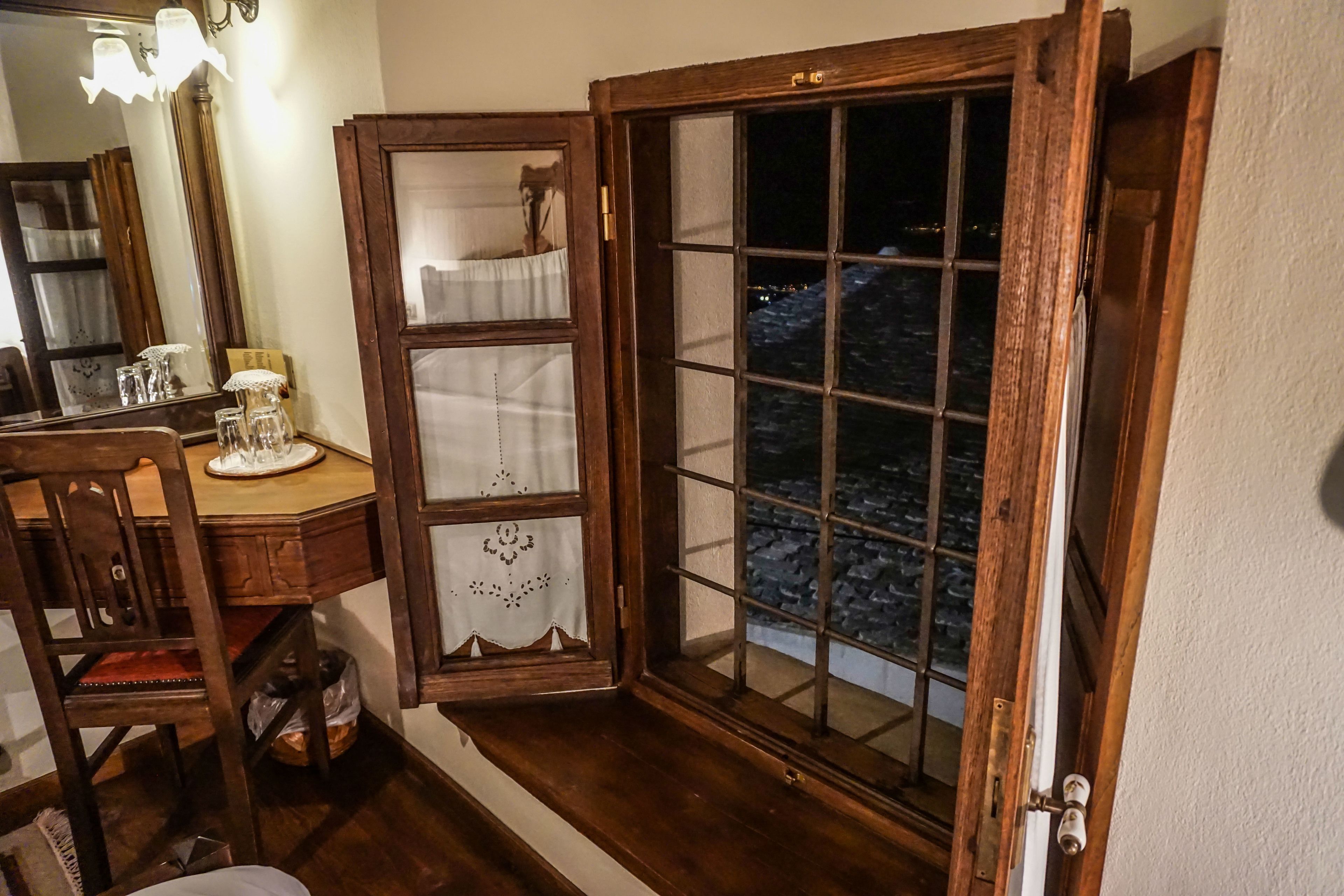 A room with a wooden desk, a chair, a window with a view of a cityscape at night, and a mirror with a light fixture.