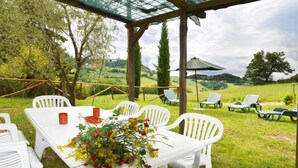 Un espacio al aire libre con sillas y mesas de plástico blanco, una pérgola de vidrio y vistas a un paisaje verde con árboles y colinas.