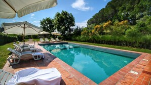 Una piscina rectangular con borde de azulejos, rodeada de sillas de descanso y sombrillas, con árboles y vegetación de fondo.