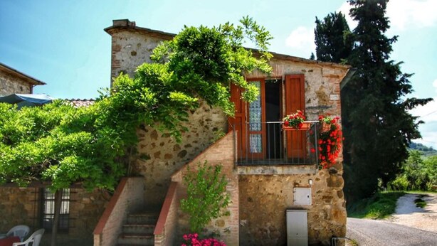Un edificio de piedra con una escalera, un pequeño balcón con flores rojas y un jardín con flores rosadas.