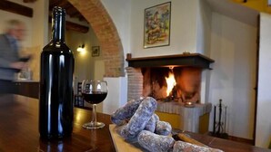 Una botella de vino, una copa de vino tinto y varios embutidos sobre una mesa de madera en una habitación con chimenea de ladrillo.