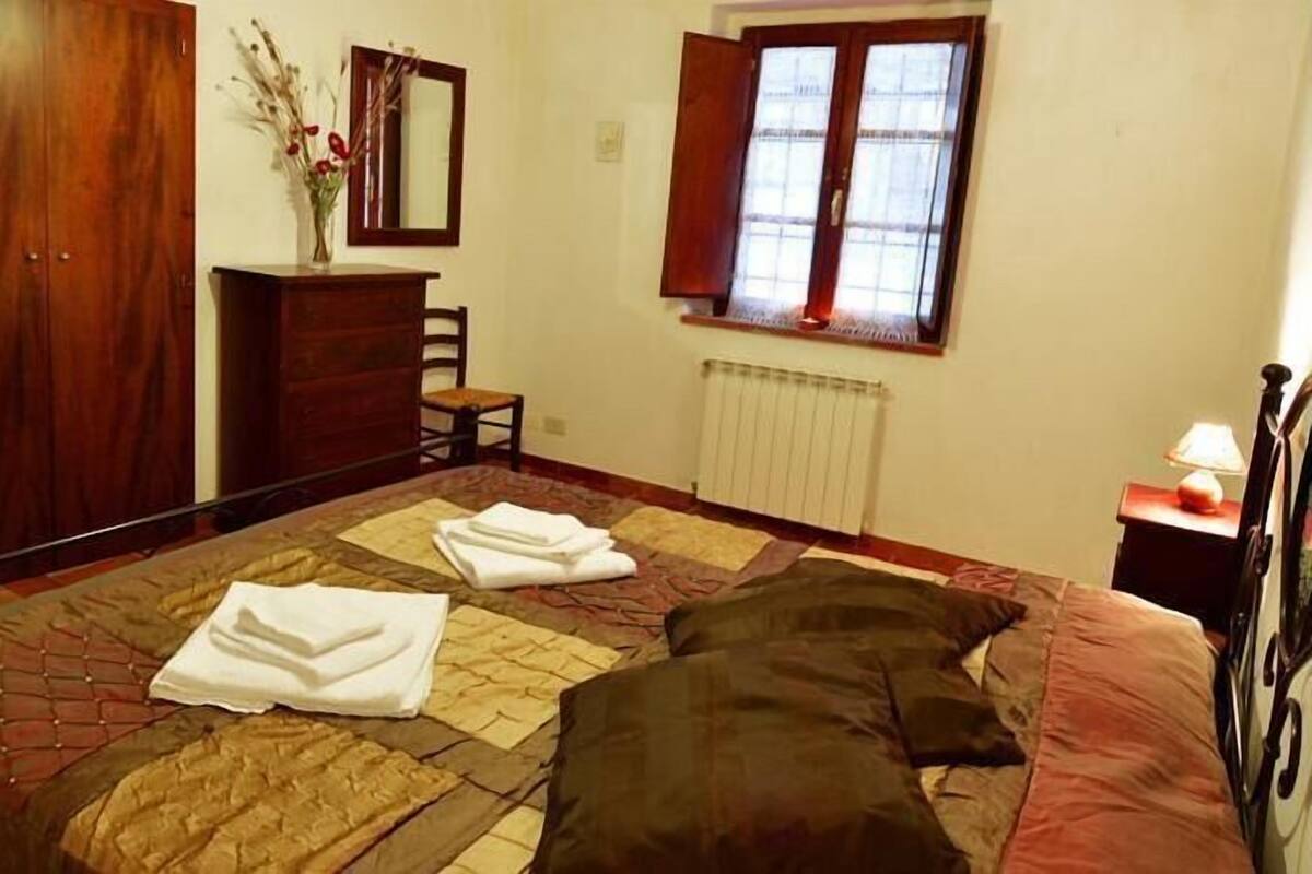 Un dormitori amb un llit gran, un armari de fusta, una petarrera, una cadira, un radiador i una finestra amb cortines.