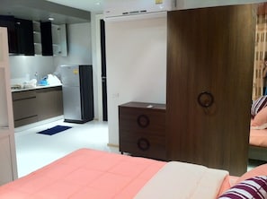 Quarto de hotel com cama, guarda-roupa de madeira, cozinha compacta com frigorífico e um tapete azul no chão.