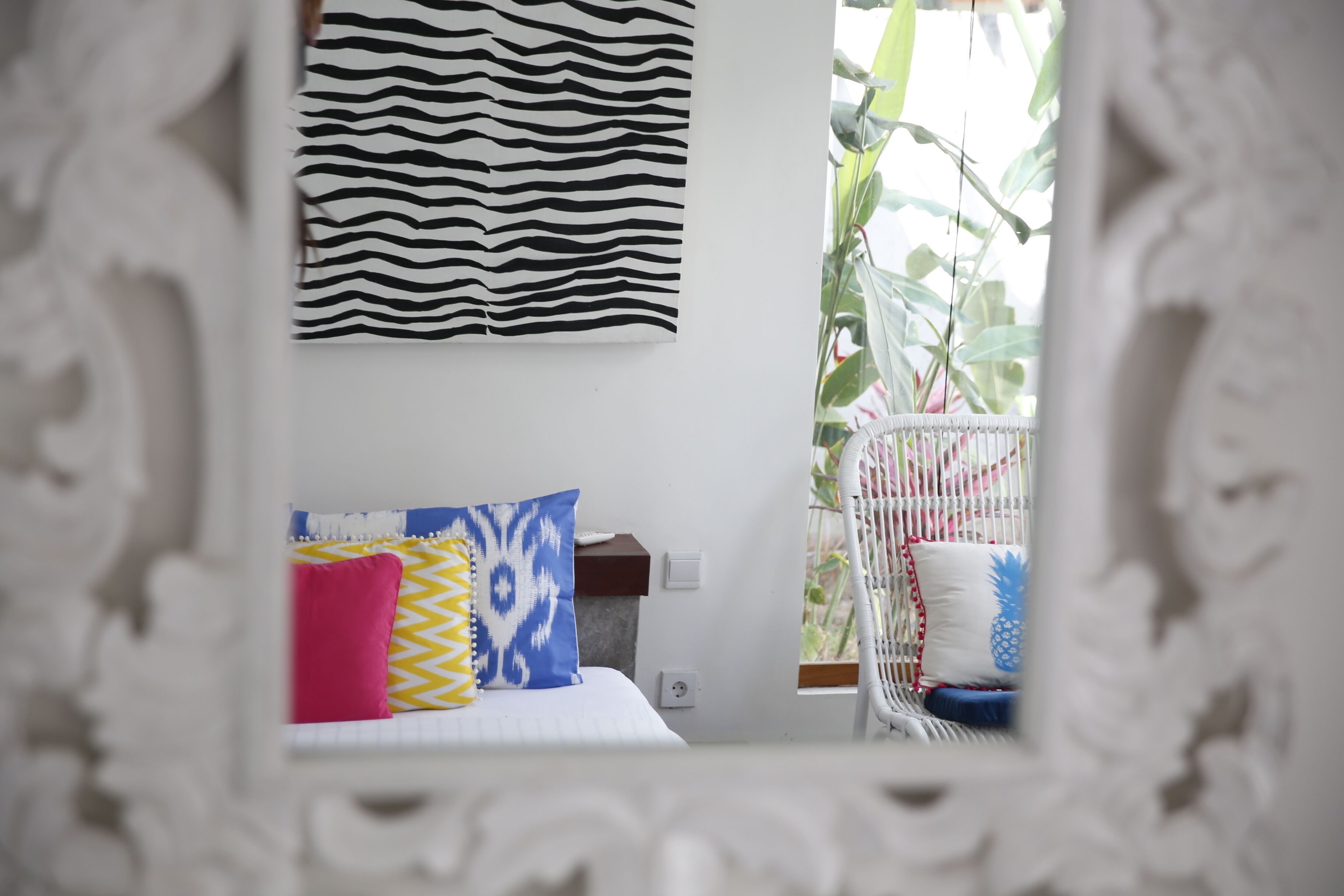 A bed with patterned pillows, a white wall, and a window with a view of greenery.
