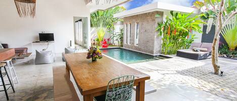 A modern outdoor dining area with a pool, surrounded by lush greenery and a clear blue sky.