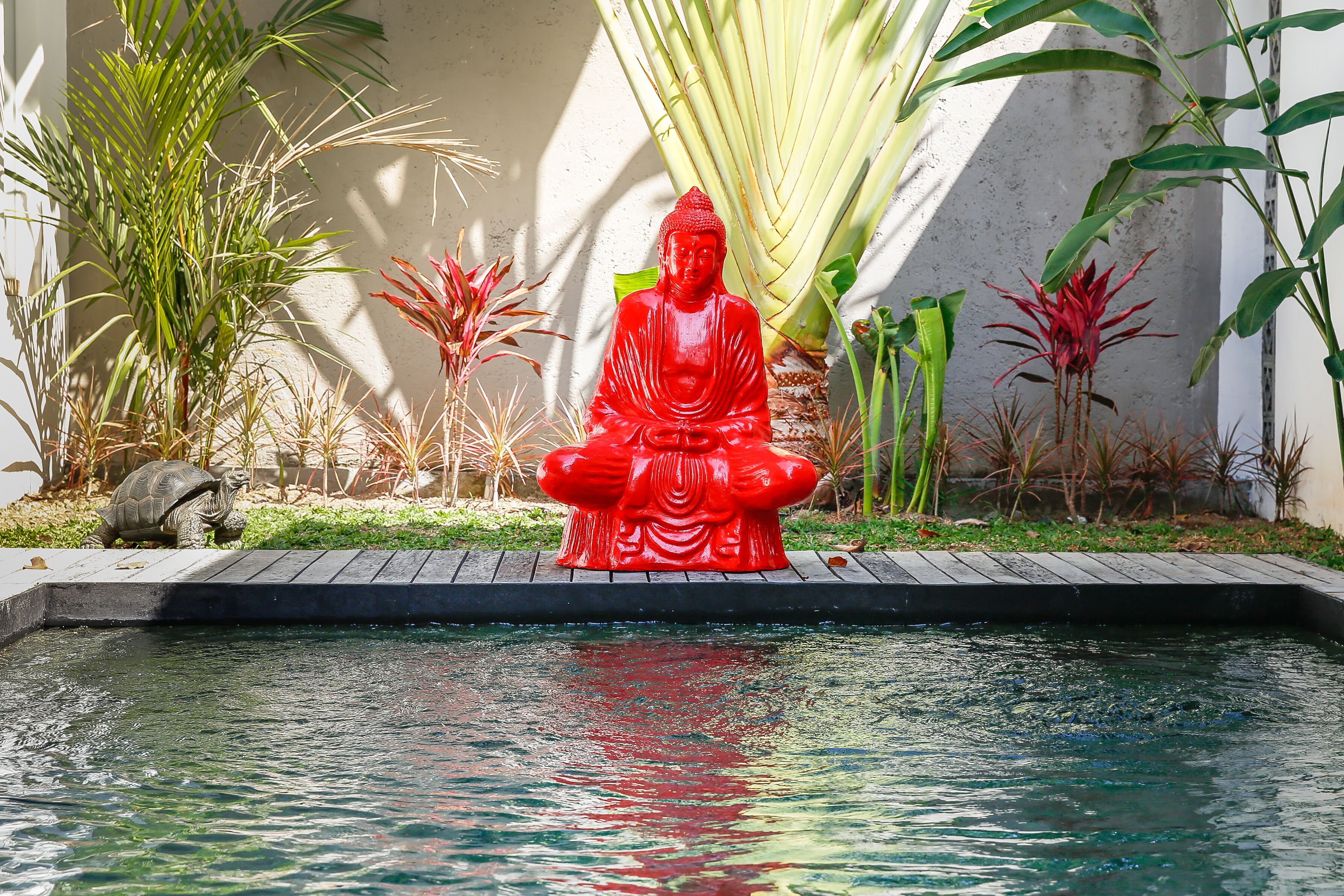 A red Buddha statue by a pool with surrounding greenery and a turtle.