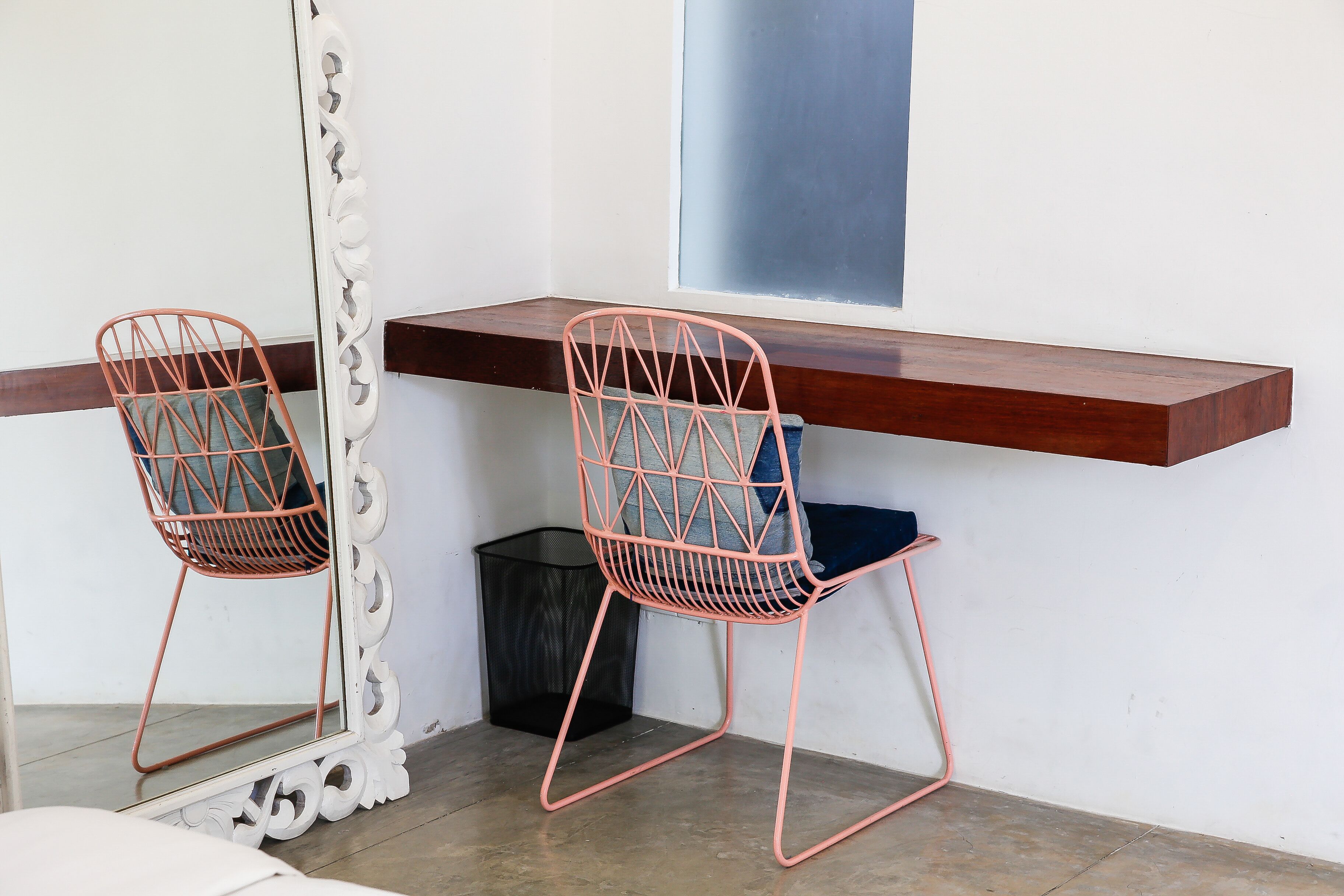 A room with a wooden bench, two wireframe chairs, a mirror, and a trash bin.