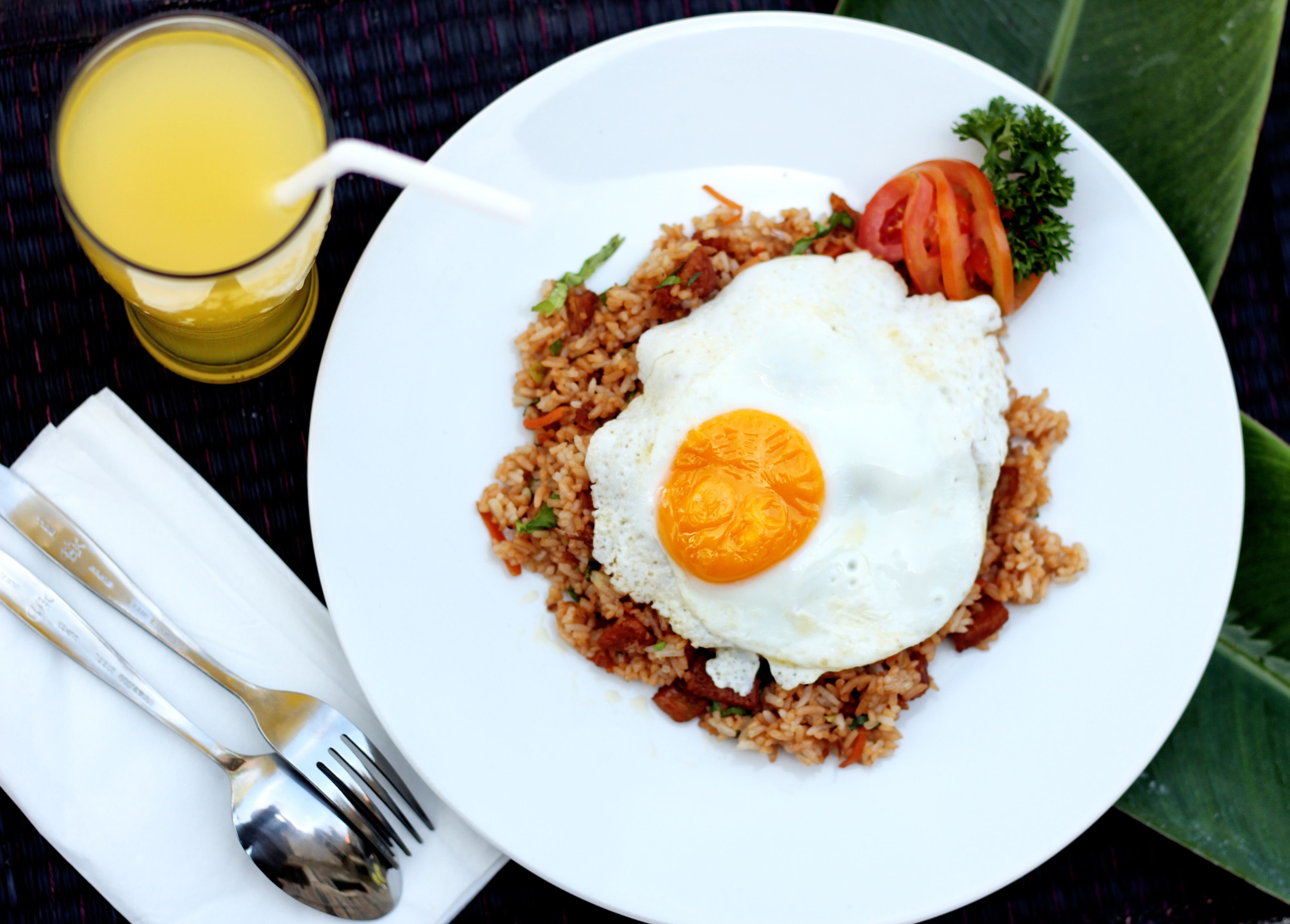 A plate of fried rice with an egg, a slice of tomato, and a sprig of parsley, accompanied by a glass of orange juice and cutlery on a table.