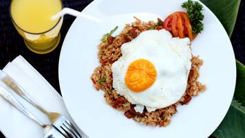 A plate of fried rice with an egg, a slice of tomato, and a sprig of parsley, accompanied by a glass of orange juice and cutlery on a table.